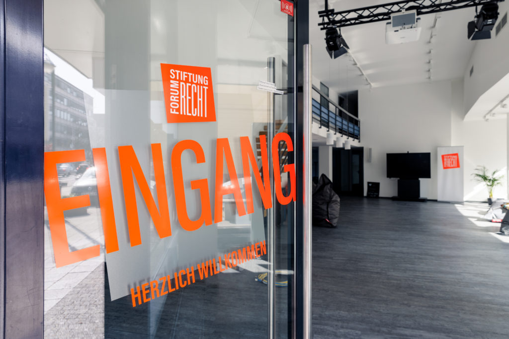 Offene Eingangstüre mit orangenem Schriftzug "Eingang". Blick auf das innere des Veranstaltungsraums der Stiftung Forum Recht in Karlsruhe.