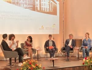 Foto Podium mit einem Stuhlhalbkreis. Auf der Wand Projektion mit Bildern eines Hauses, im Vordergrund zwei Blumensträuße. Auf dem Stuhlkreis sitzen sechs Personen. 2. v.l. Muheterem Aras (Landtagspräsidentin Baden-Württemberg). 2. v.r. Dr. Frank Mentrup (Oberbürgermeister Karlsruhe)