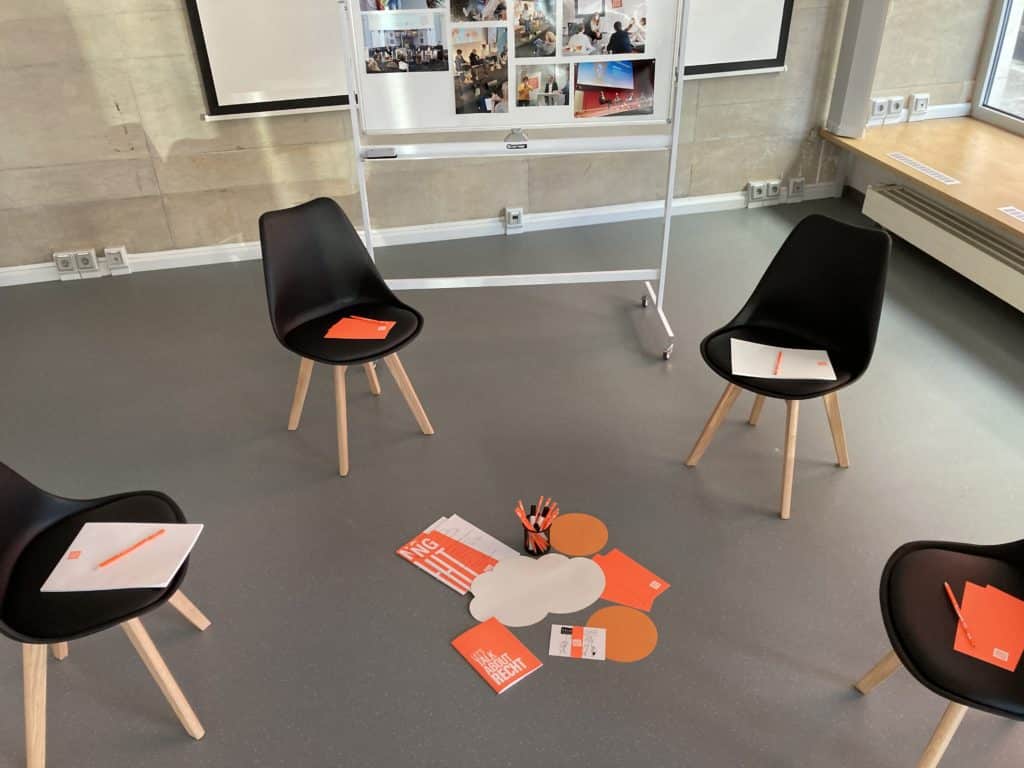 Veranstaltungsraum der Stiftung Forum Recht in Leipzig mit vier schwarzen Stühlen und neonorangem Workshop-Materialien. Im Hintergrund steht ein Whiteboard mit angepinnten Fotografien vergangener Veranstaltungen.