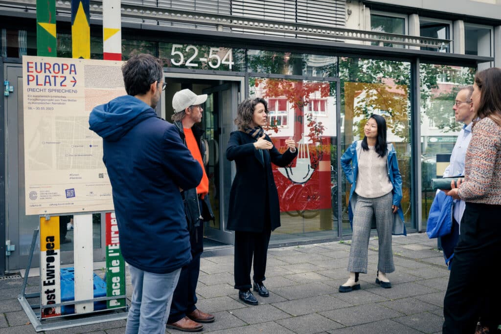 Gruppe von Menschen vor einem Gebäude und großem Plakat des Projekts Europaplatz - Recht sprechen. Eine Frau erzählt mit Handgeste.