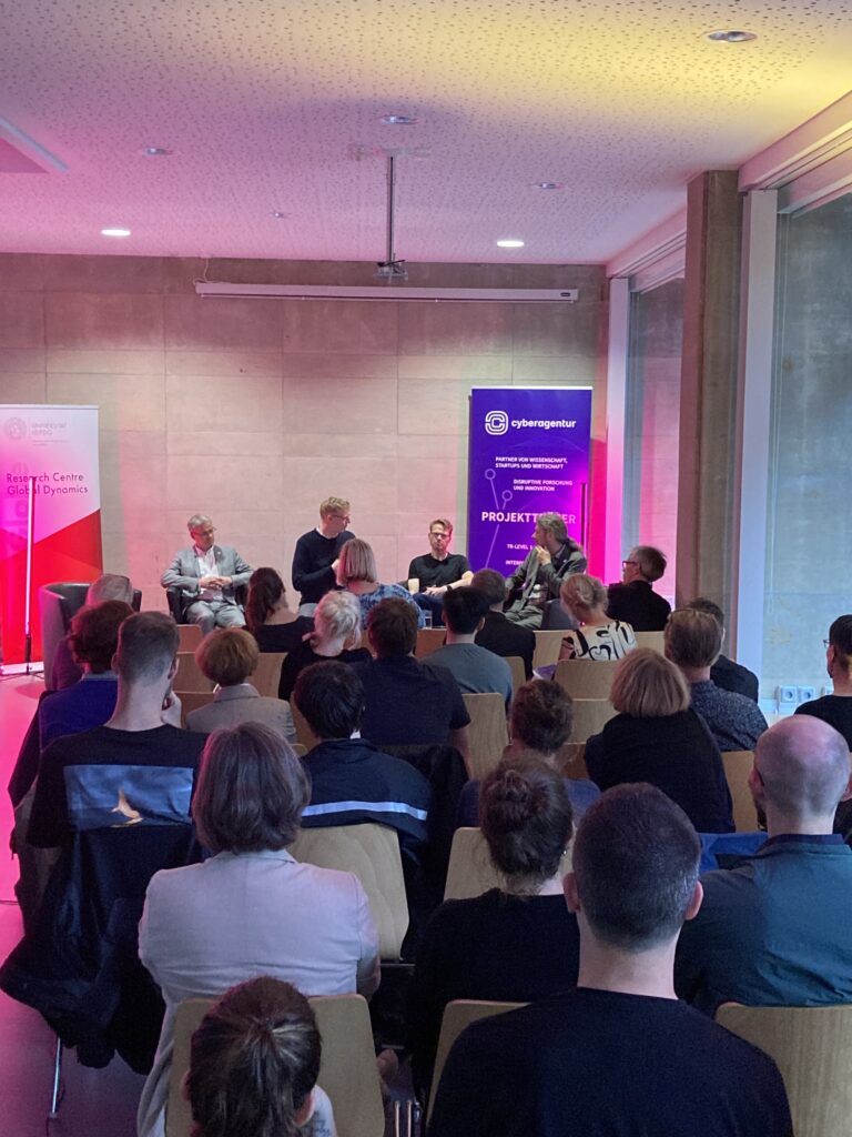 Foto des Veranstaltungssaals der Stiftung Forum Recht Leipzig. Publikum und am Ende des Raums ein Podium mit vier Referenten
