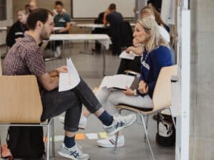 Workshopgruppe auf Stühlen im Raum verteilt, im Vorderung unterhalten sich ein Mann und eine Frau, sie halten Papiere in der Hand, auf einem Papier ist die Aufschrift "Recht" zu erkennen