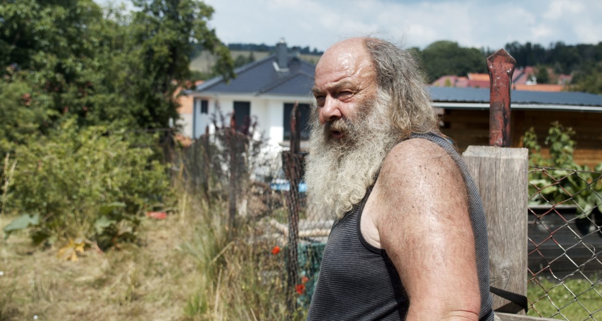 Foto. Älterer Herr mit langem Bart im Seitenprofil vor einem Gartenzaun