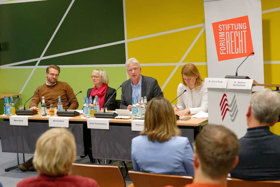 Foto. Vom Publikum aus sind vier Personen fotografiert die an einem langen Tisch mit Microfonen sitzen