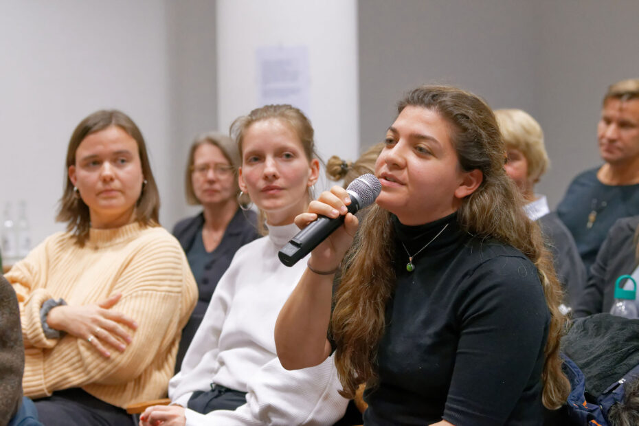 Foto: Publikum sitzend. Eine junge Frau stellt mit Mikrofon eine Frage