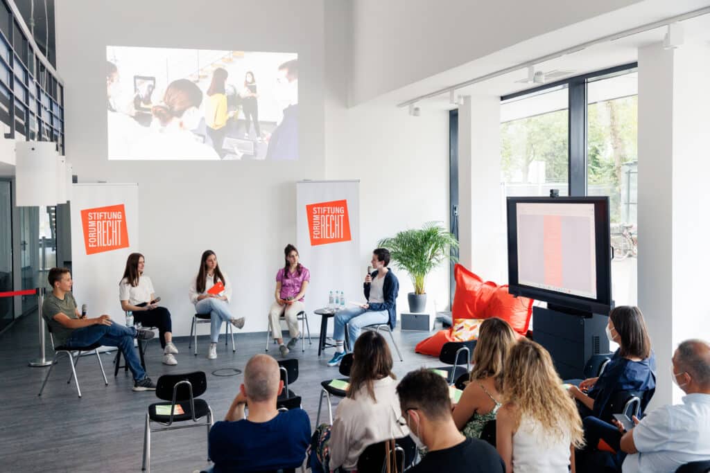 Fünf junge Menschen sitzen im Halbkreis im Gespräch vor dem Publikum in den Stiftungsräumen Karlsruhe, an die Wand wird ein Foto gebeamt