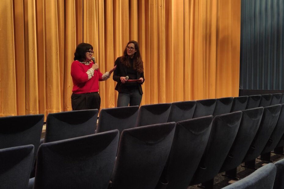 Foto. Zwei Frauen stehen vor einem goldenen Kinovorhang. Im Vordergrund Sitzreihen