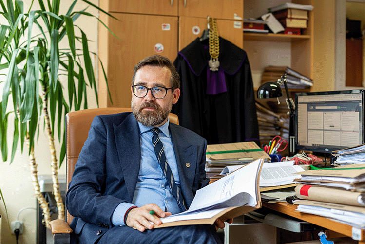 Foto eines Mannes, der im Anzug vor einem beladenen Schreibtisch sitzt und Dokumente in seinen Händen hält- Im Hintergrund hängt seine Richterrobe und -kette.