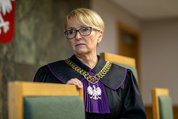 Foto einer Richterin in schwarz-lila Robe, die durch ihre Brille etwas verbittert an der Kamera vorbeischaut. Sie befindet sich in einem Gerichtssaal und trägt eine große, goldene Kette mit polnischem Adlerwappen.