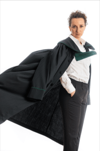Foto einer Rechtswanwältin, die mit nach links wehender Anwältinnenrobe hüfthoch vor weißem Hintergrund steht und ernst in die Kamera blickt.