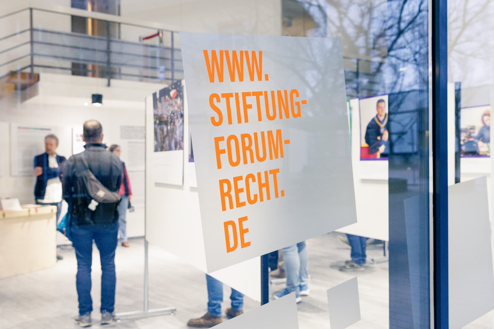 Blick auf das Schaufenster der Stiftung in Karlsruhe, darauf ist groß die Webadresse der Stiftung in orange gedruckt, im Innenraum sieht man die Fotoausstellung Sprawiedliwość - Gerechtigkeit und Besuchende