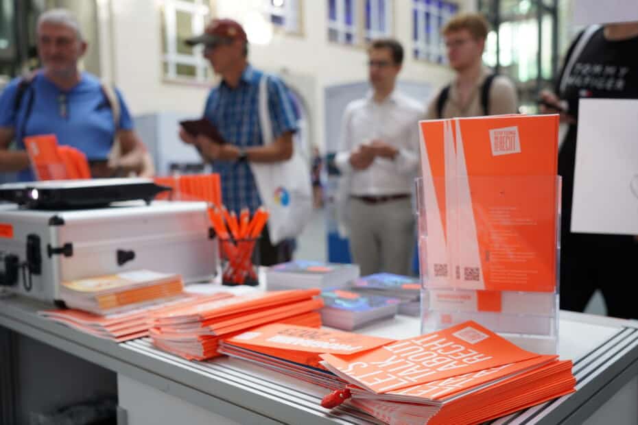 Blick vom Info-Stand der Stiftung Forum Recht, mit neon-orangenen Info-Broschüren, die Standbesucher sind unscharf im Hintergrund