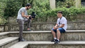 Ein Junger Mann filmt stehend einen Jungen, der seitlich auf einer Treppe sitzt. Beide Personen sind im Profil zusehen. Der sitzende Jugendliche trägt einen Ohring und hat kurze haare. Der filmende Mann ist vom Alter her vielleicht Studierender.