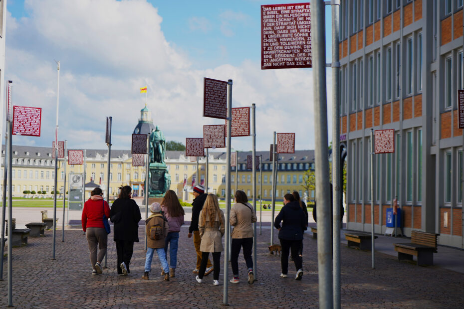 Menschen verschiedenen Alters laufen über den Karlsruher Platz für Menschenrecht. Im Hintergrund sieht man das Stadtschloss.