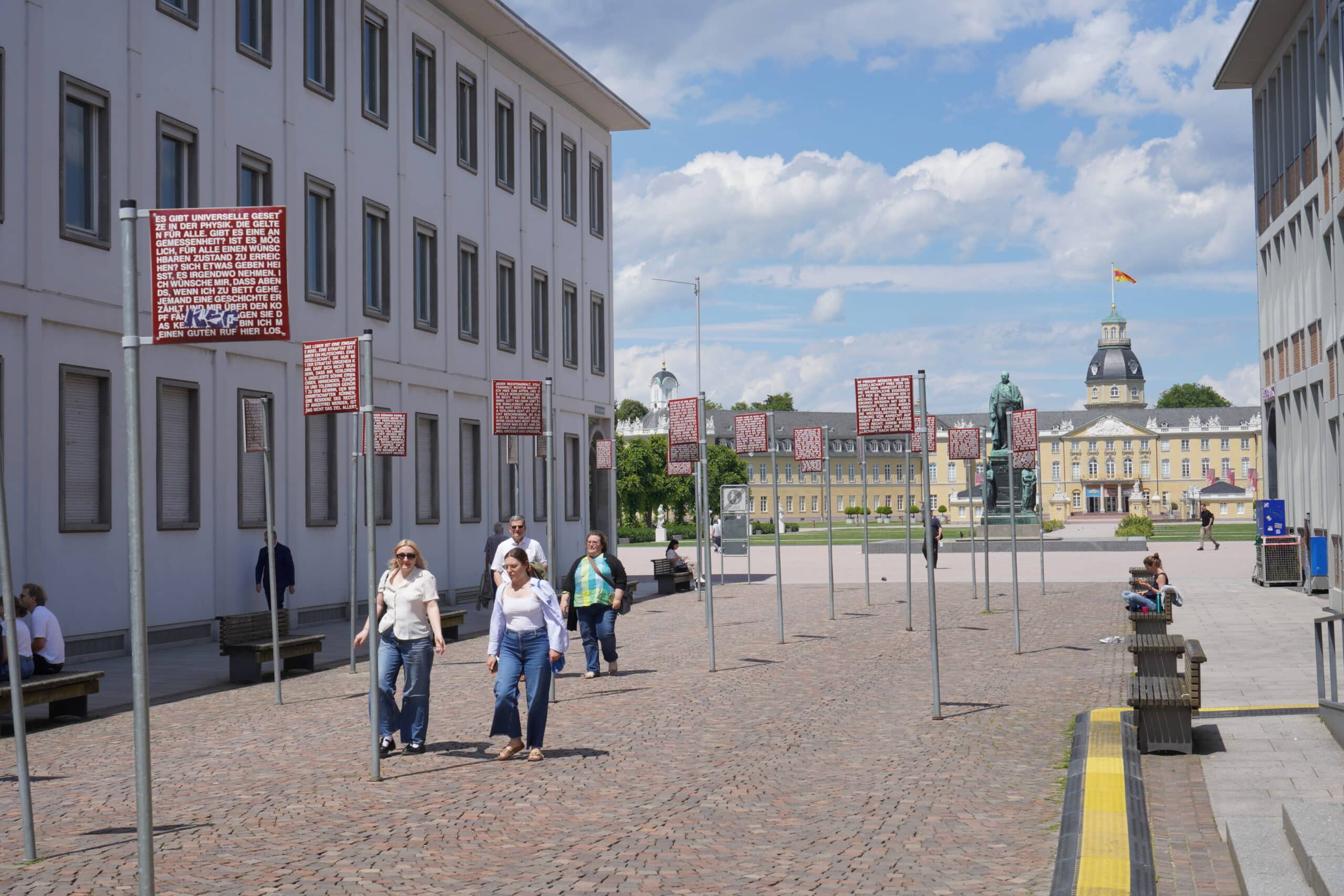 Blick auf die Schilderbehangene Pflastersteinstraße, die zum Karlsruher Schloss führt. Der Himmel ist leicht bewölkt uns es sind ein paar Menschen auf der Straße.