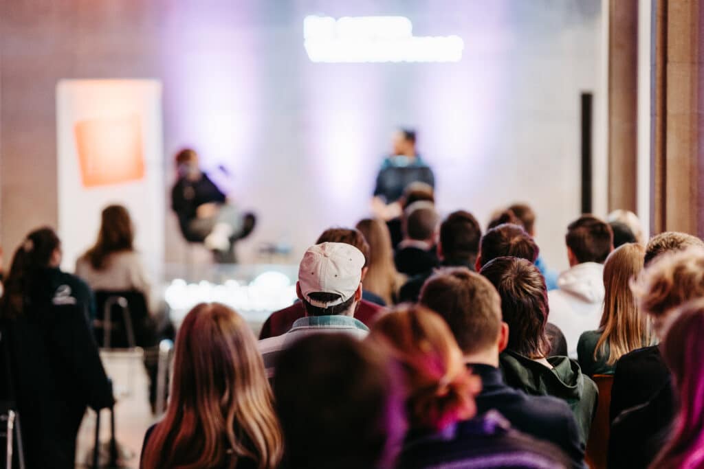 Foto mit Unschärfen, Blick auf die Live-Podcast-Bühne in der Stiftung Forum Recht. Im Vordergrund das bunte Publikum, sitzend und von hinten aufgenommen. Im unscharfen Hintergrund Moderatorin Rabea Schloz im Gespräch mit dem Journalisten Arne Semsrott.