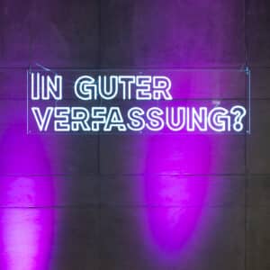 Ein neonblaues Leuchtschild mit dem Schriftzug "In guter Verfassung" hängt vor einer lila beleuchteten Wand.