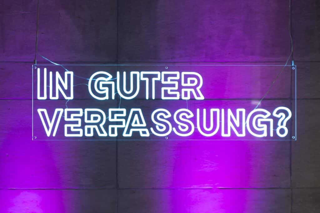 Ein Leuchtschild mit dem neonblauen Schriftzug "In guter Verfassung" hängt vor lila beleuchteten Wänden.