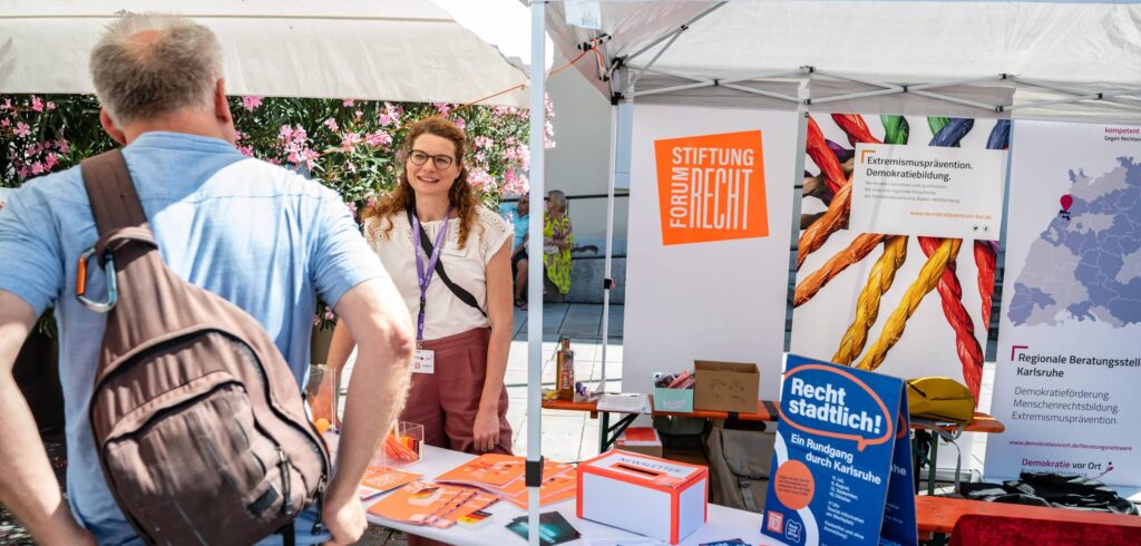 Eine Mitarbeiterin der Stiftung Forum Recht steht an einem überdachten Informationsstand und ist im Gespräch mit einem interessierten Mann,