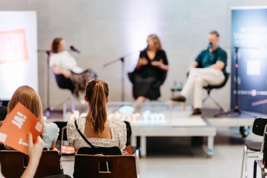 Im Vordergrund zwei junge Zuschauerinnen, die von hinten auf Stühlen sitzend fotografiert wurden. Im Hintergrund unscharf die Podcast-Live-Event Bühne mit der Moderatorin und zwei Gästen.