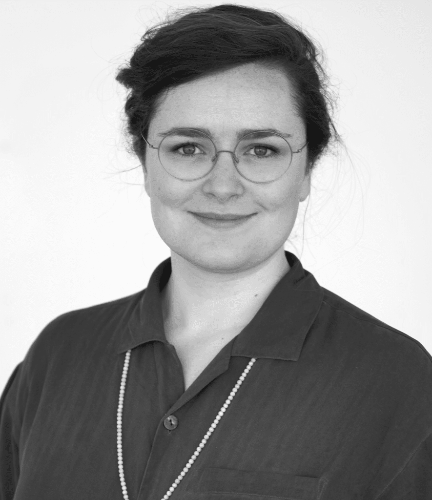 Portrait of a woman with round glasses. She smiles at the camera. Her hair is pinned back at the back of her head.