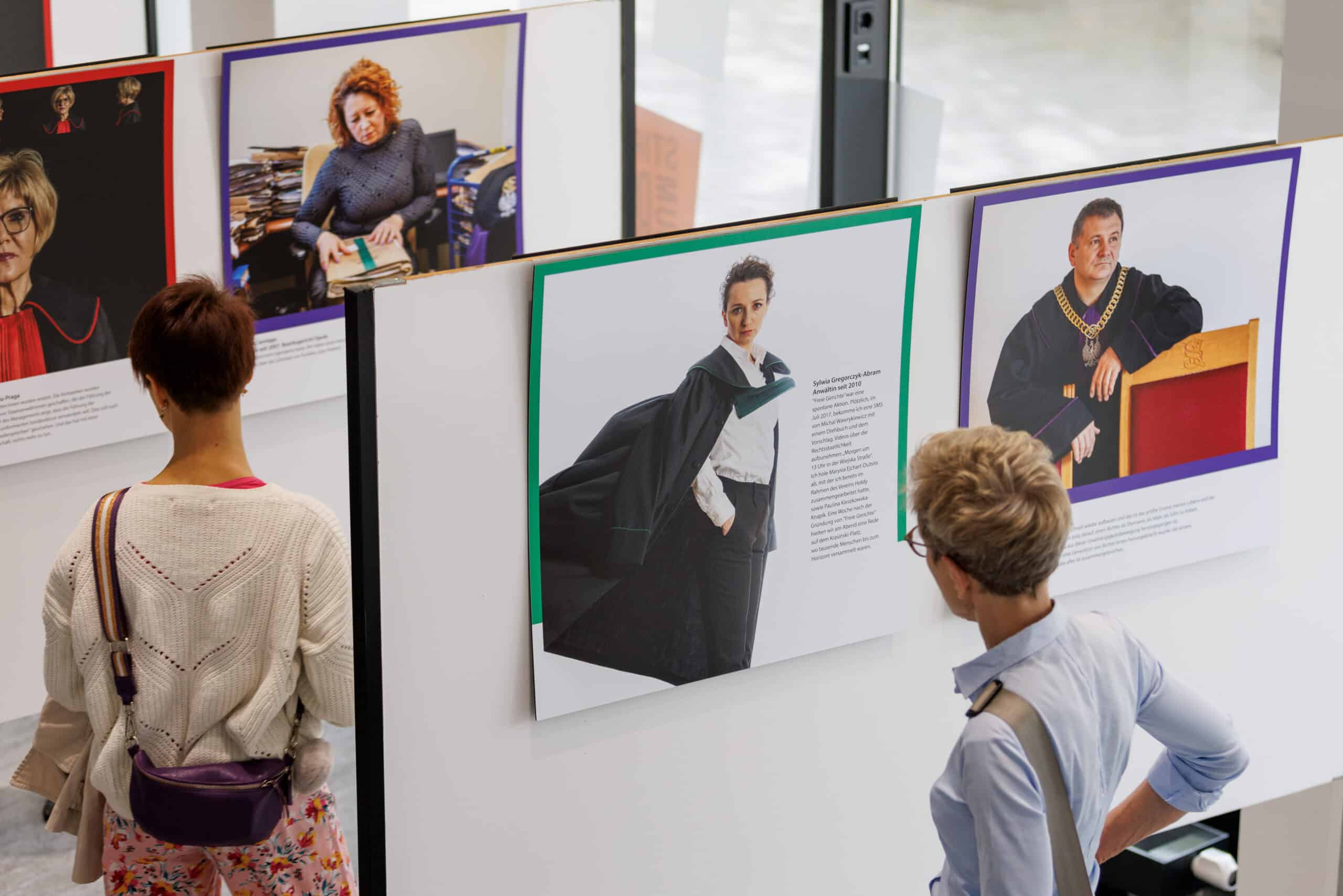 Zwei Personen betrachten Fotos von Richter:innen und Staatsanwätlt:innen, die in einer Ausstellung hängen.