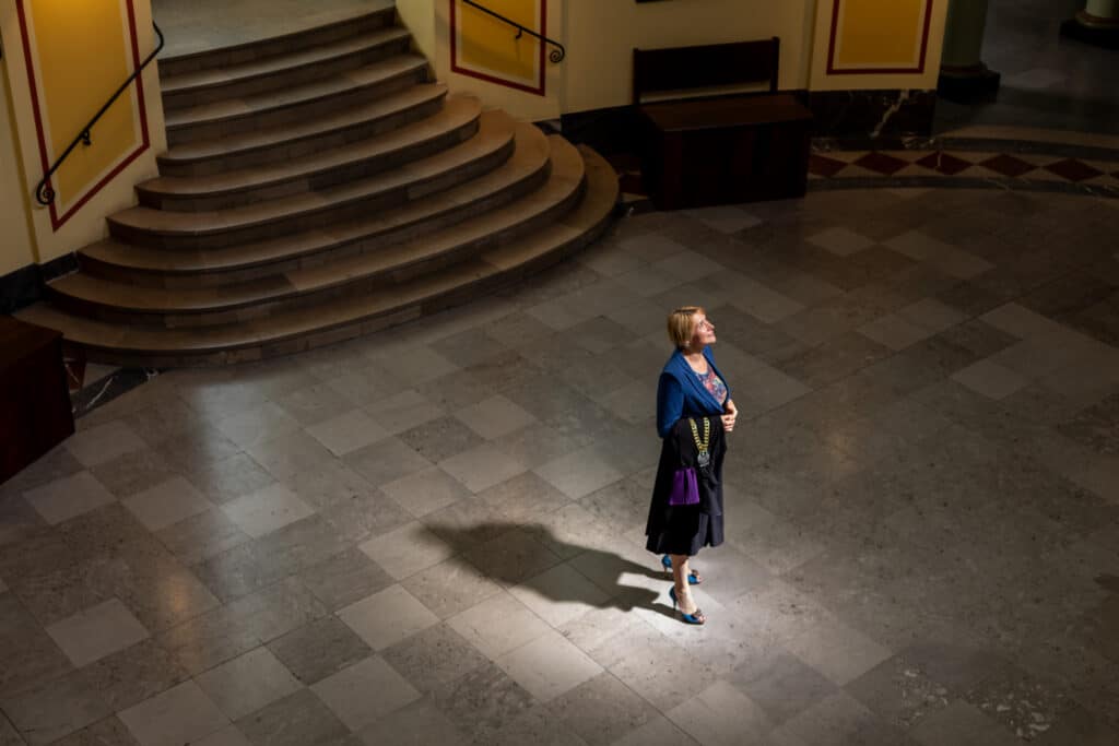 Eine blonde Frau in lila Sakko steht in der Eingangshalle eines Gerichtsgebäudes und blickt freudig in das auf die herabfallende Sonnenlicht. Sie hat die schwarz-lila Richterinnenrobe mit goldener Kette und Adlerwappen über ihren rechten Unterarm geworfen.