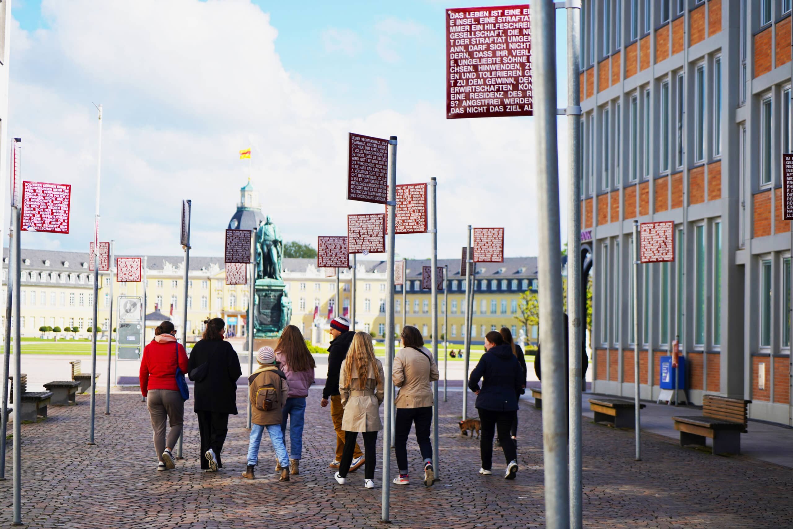 Eine Gruppe von Erwachsenen und ein Kind laufen vor dem Karlsruher Schloss über einen Pflastersteinplatz. Sie laufen eine kupferne Statue zu.