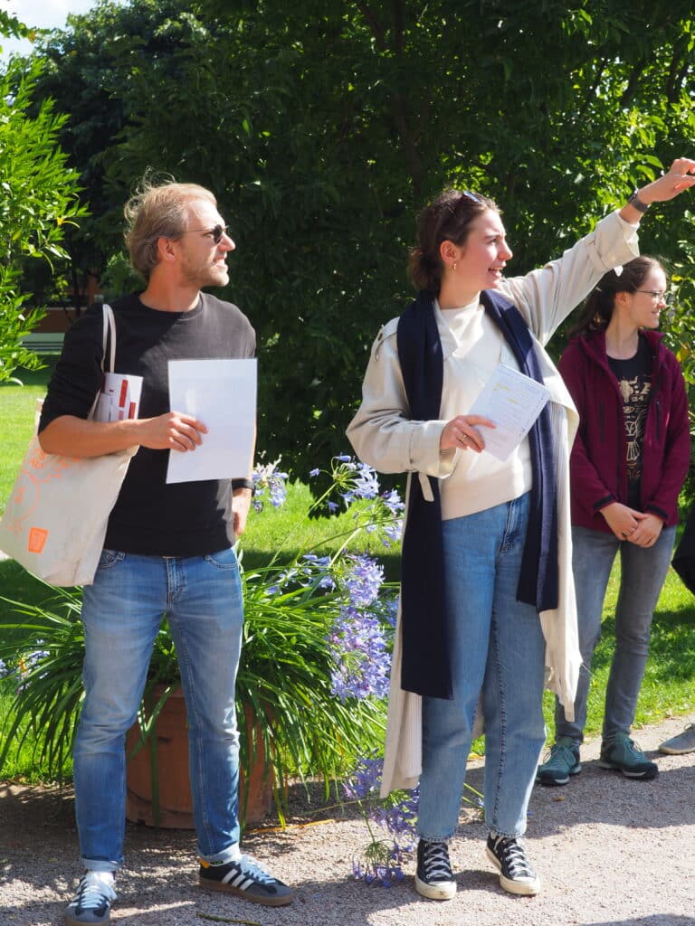 Zwei Mitarbeiter:innen der Stiftung stehen leichter Herbstkleidung in einem grünen Park. Die Sonne scheint.