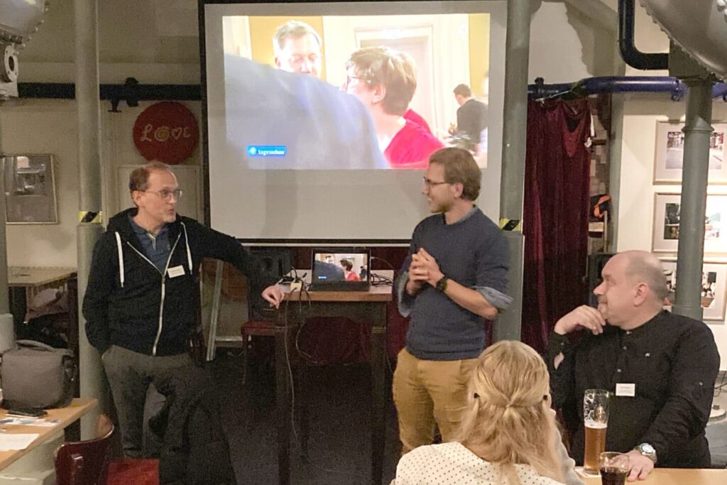 Drei Männer und eine Frau stehen und sitzen vor einer kleinen Leinwand, die in einer Gaststätte aufgebaut ist.