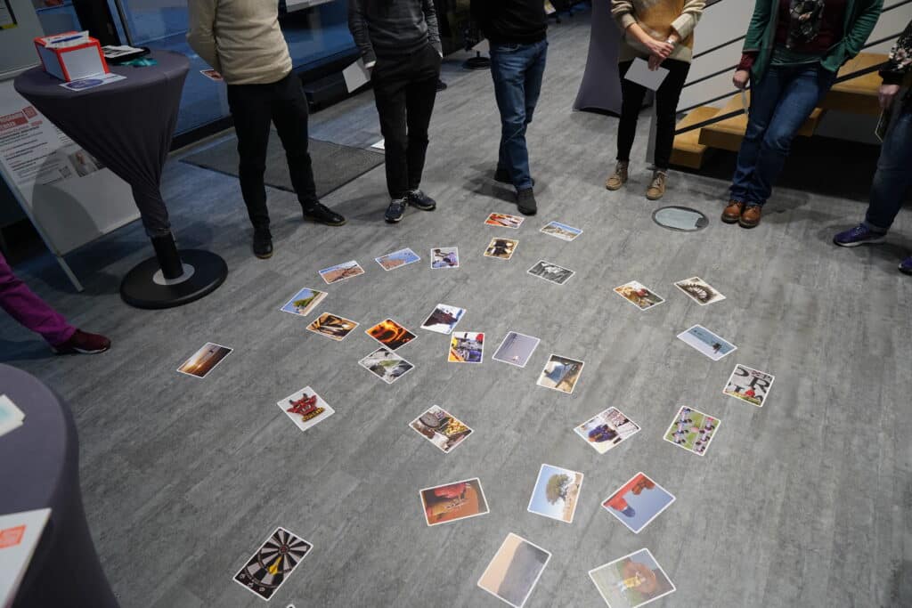 Szene aus einem Bildungsworkshop. Verschiedene Fotografien liegen auf grauem Boden in den Räumen der Stiftung Forum Recht. Darum herum stehen ein paar Teilnehmer:innen nachdenklich