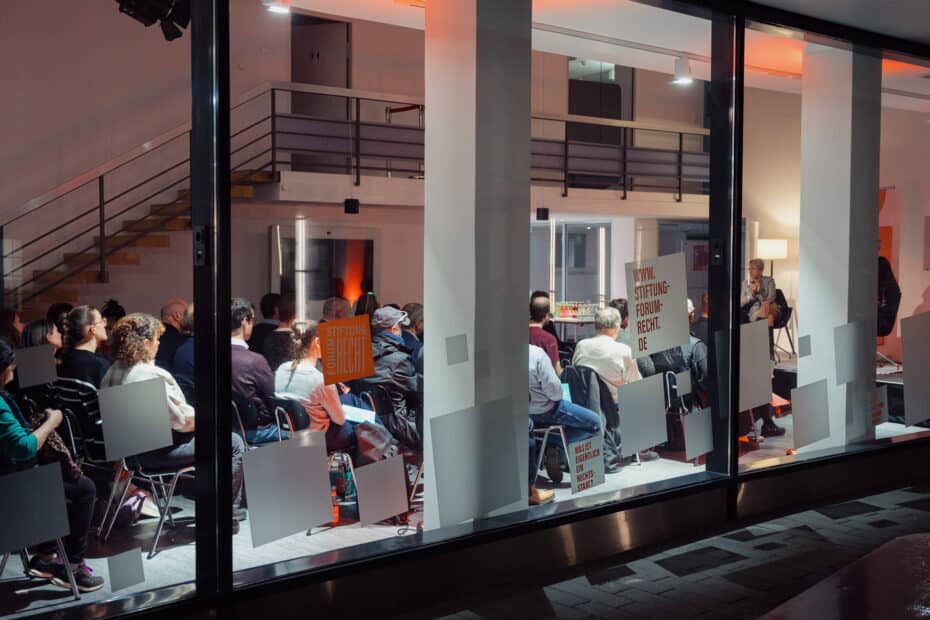 Veranstaltungsraum von außen durch große Glasscheiben fotografiert - mit Publikum, das auf Stühlen sitzt.