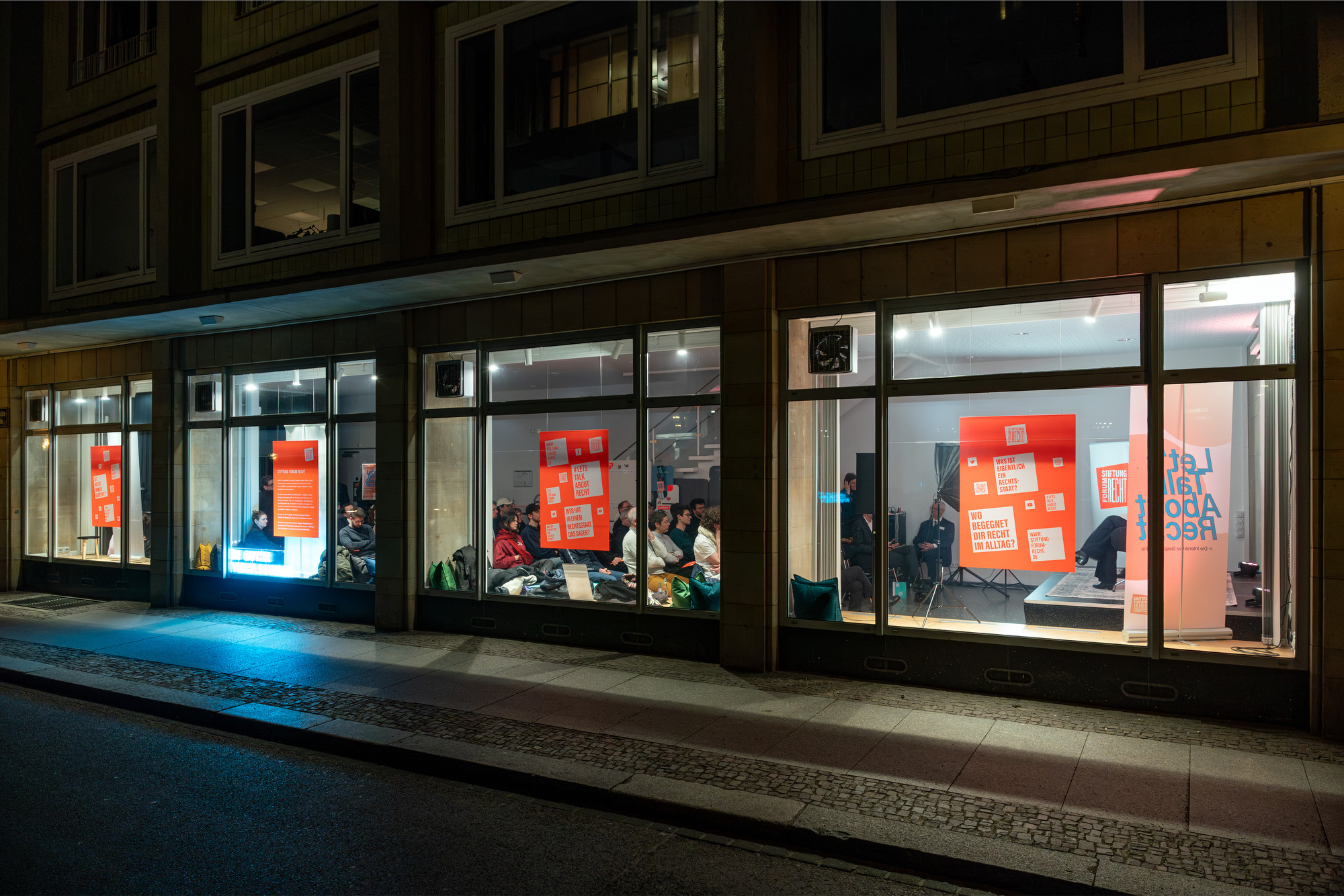 Blick durch eine eine ebenerdige Fensterfront in einen Veranstaltungsraum, in dem Publikum in Sitzreihen sitzt. In den Fenstern hängen große orangene Plakate der Stiftung Forum Recht.