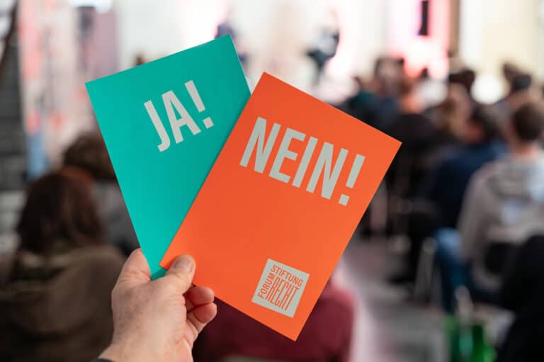 Eine Hand hält zwei Karten: links eine grüne Karte mit Ja, rechts eine rote Karte mit Nein. Im Hintergrund Menschen in einem Veranstaltungsraum.