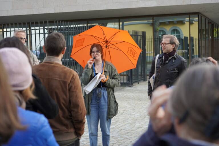 Eine junge Frau unter einem orangen Stiftungsregenschirm spricht zu den Teilnehmenden des Stadtrundgangs.