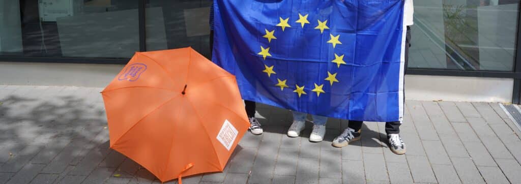 Europaflagge aus dunklem Blau mit Sternkreis in der Mitte, daneben ein knallig oranger Schirm