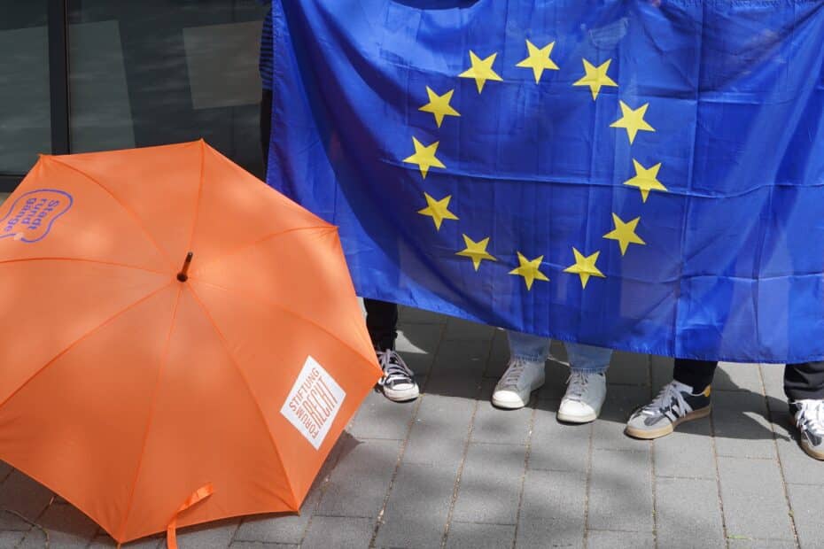 Europaflagge aus dunklem Blau mit Sternkreis in der Mitte, daneben ein knallig oranger Schirm