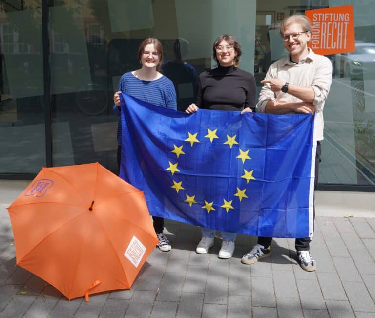 Drei Personen, zwei junge Frauen und ein Mann vor dem Stiftungsgebäude mit einer Europaflagge und einem Regenschirm in knalligem Orange.