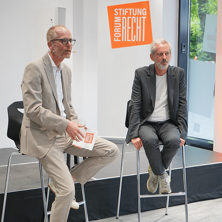 Zwei Männer auf einem Podium im Gespräch zum Publikum gewandt.