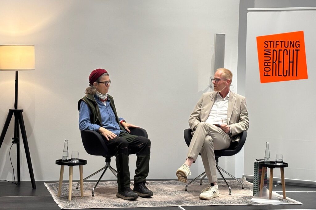 Ein mann in elegantem hellen Zweiteiler sitzt einer Frau mit Wollmütze auf dem Podium der Stiftung gegenüber und sie sind ins Gespräch vertieft.