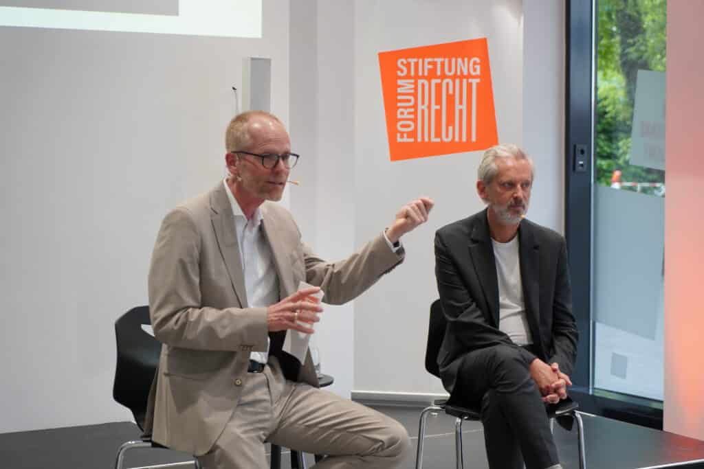 Zwei Männer auf einem Podium im Gespräch zum Publikum gewandt.