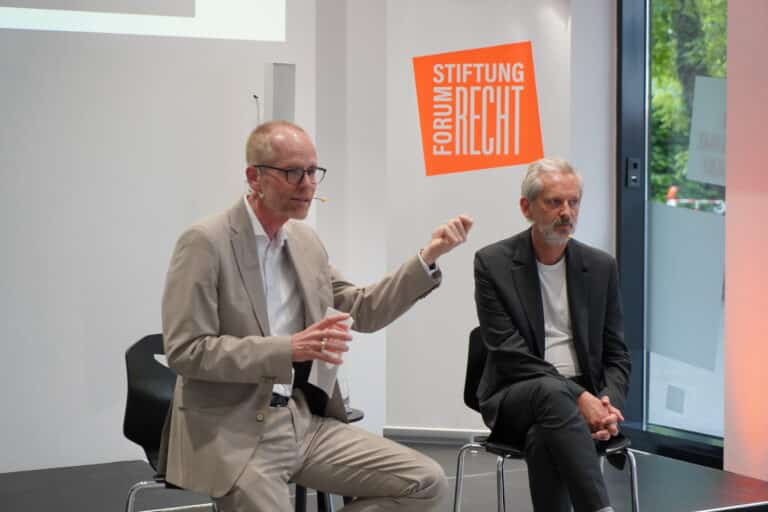 Zwei Männer auf einem Podium im Gespräch zum Publikum gewandt.