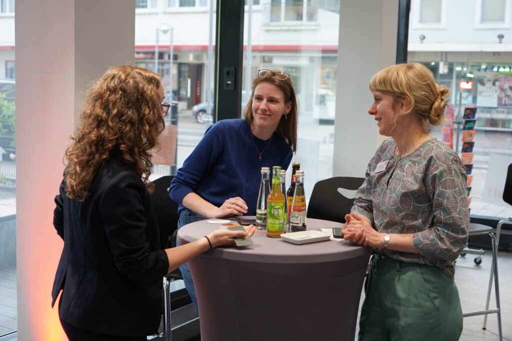 Drei junge Frauen im Gespräch an einem Stehtisch, darunter auch Anna Nübling, Programmkuratorin der Stiftung Forum Recht.