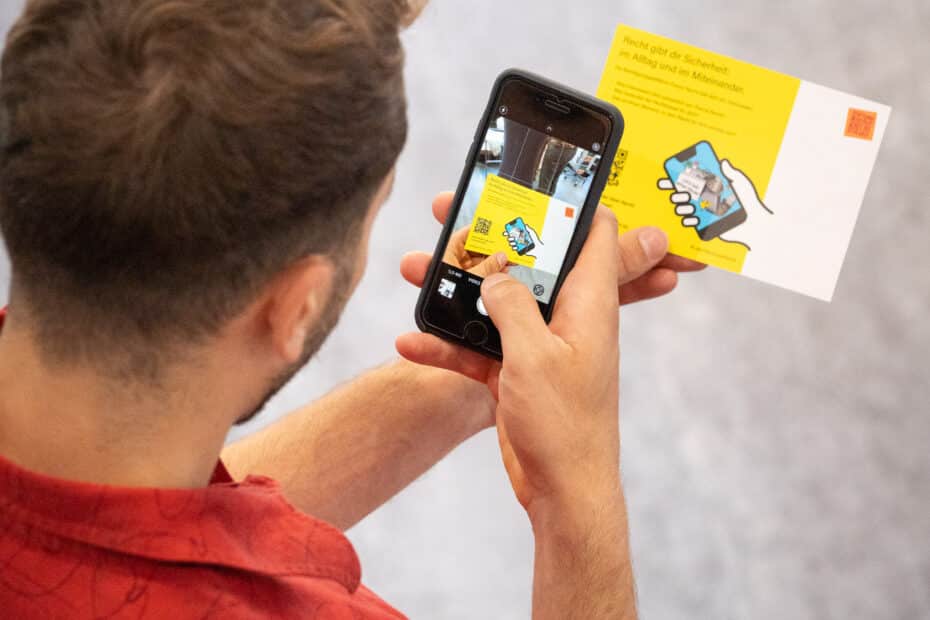 Junger Mann mit rotem Hemd wird von hinten und oben dabei fotografiert, wie er das Beteiligungstool auf dem Smartphone bedient