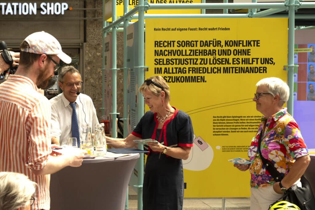 Fünf Personen stehen um einen Stehtisch. Zwei Frauen rechts schauen auf Postkarten, die sie in ihren Händen halten.