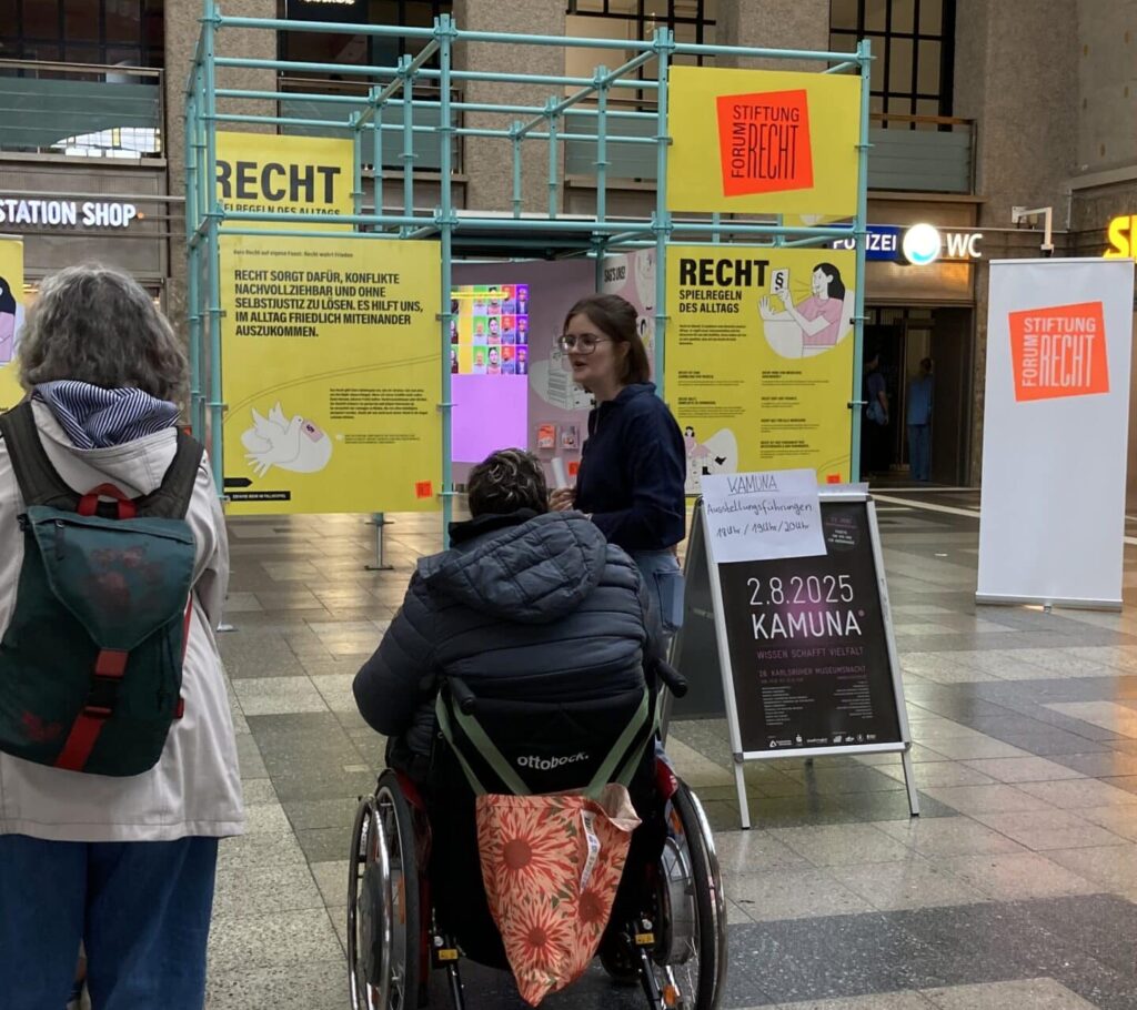 Zwei Frauen stehen, eine Frau sitzt im Rollstuhl vor einer mit Baugerüsten aufgebauten Ausstellung im Karlsruher Hauptbahnhof