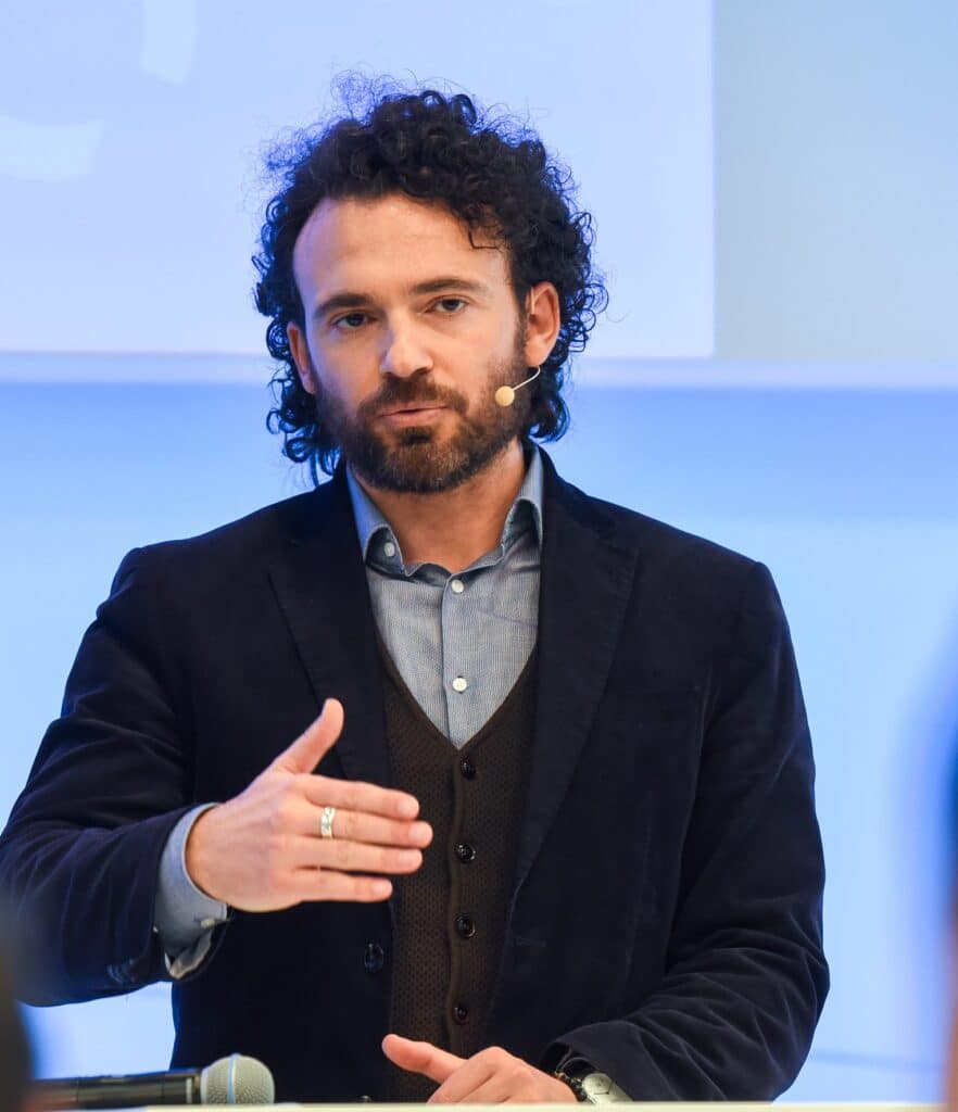 Mann mit dunklen Locken im Anzug redet und gestikuliert auf der Bühne