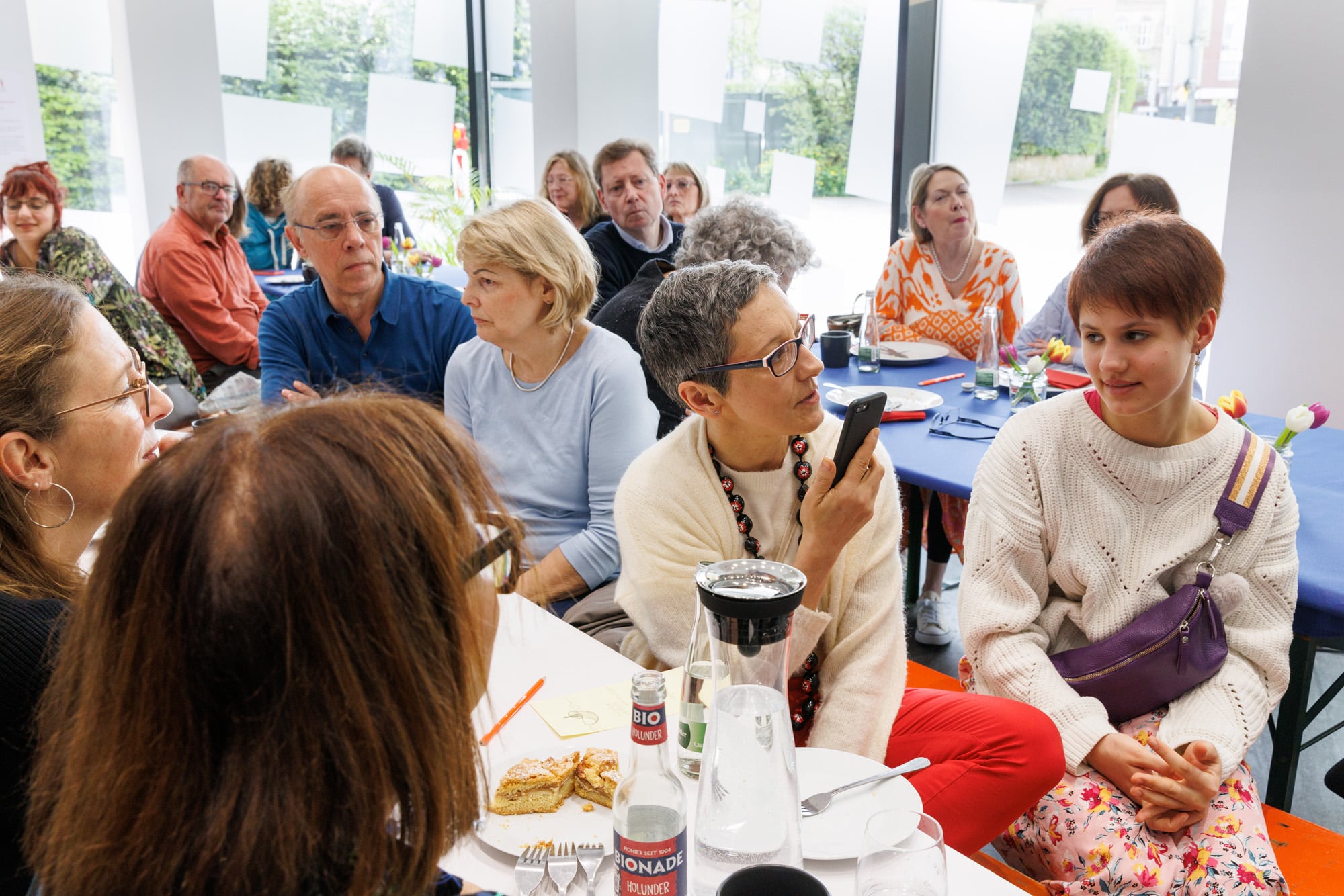 Menschen an Biertischgarnituren mit Tischdecken mit Snacks und Getränken im Gespräch.