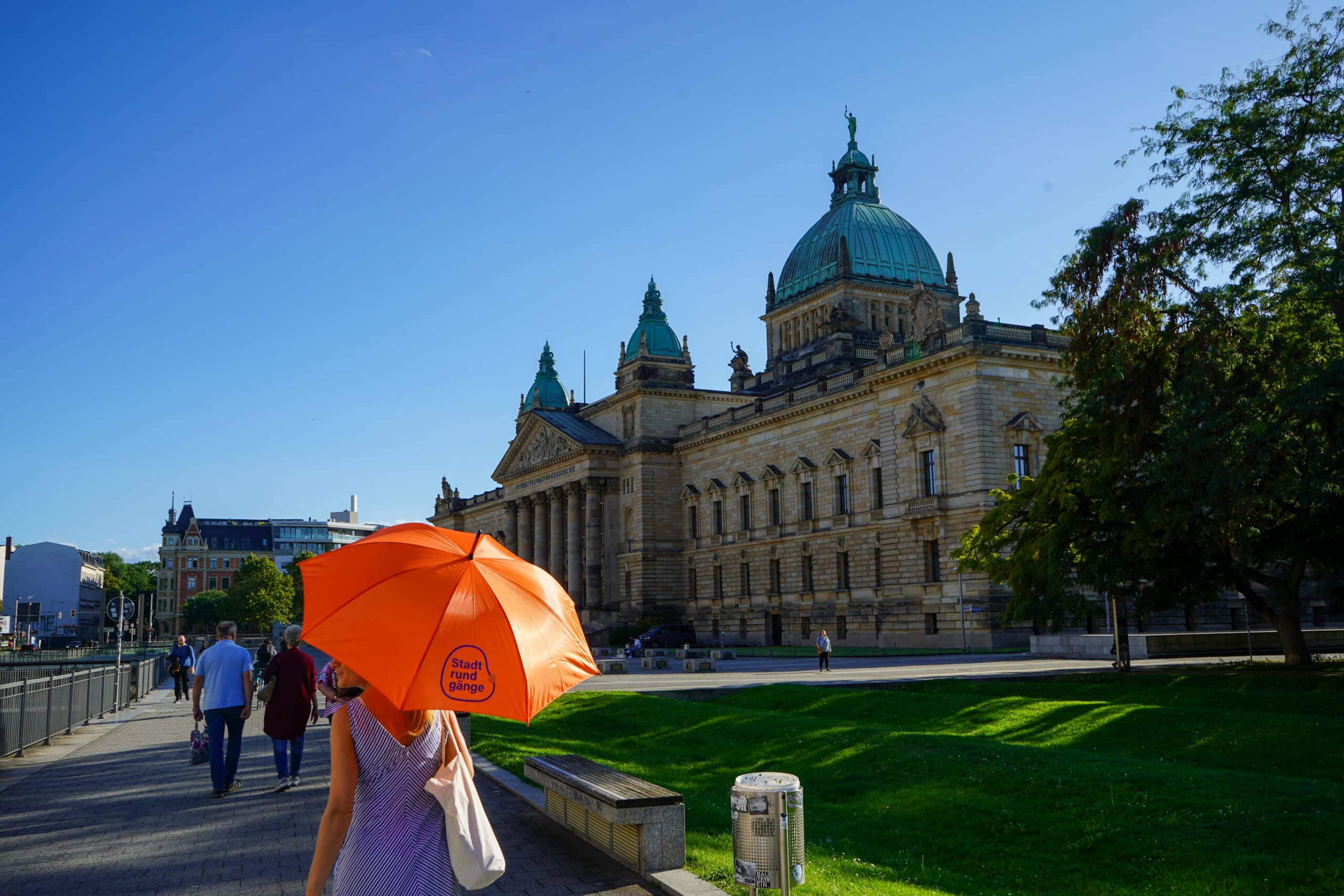 Das Bundesverwaltungsgericht; davor geht eine Frau im Sommerkleid mit einem knallorangen Schirm auf dem steht "Recht stadtlich".