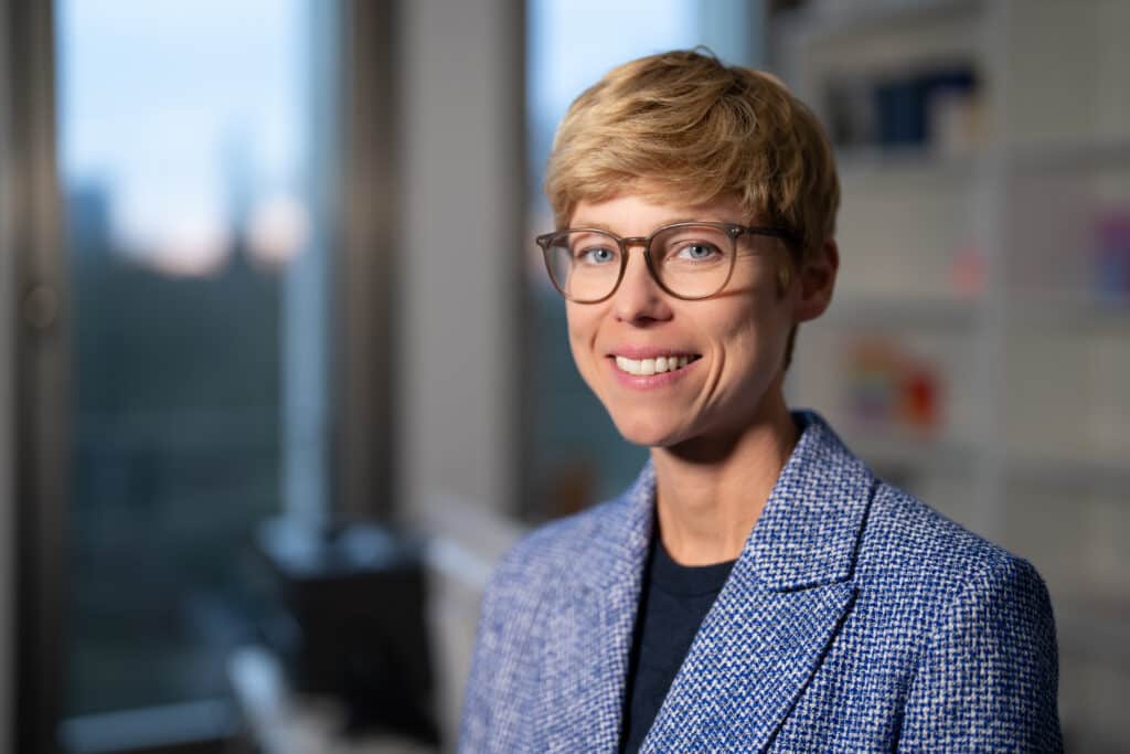 Eine junge Frau mit blonder Kurzhaarfrisur und einer Brille lächelt in die Kamera.
